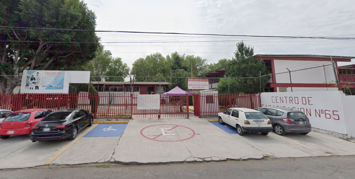 Fotografía en la que se muestra la entrada del plantel, conformada por una reja de color rojo. Del lado derecho de la reja hay un muro de color blanco, que en letras rojas dice el nombre del plantel y frente a la reja hay una rampa que da acceso a las instalaciones.
